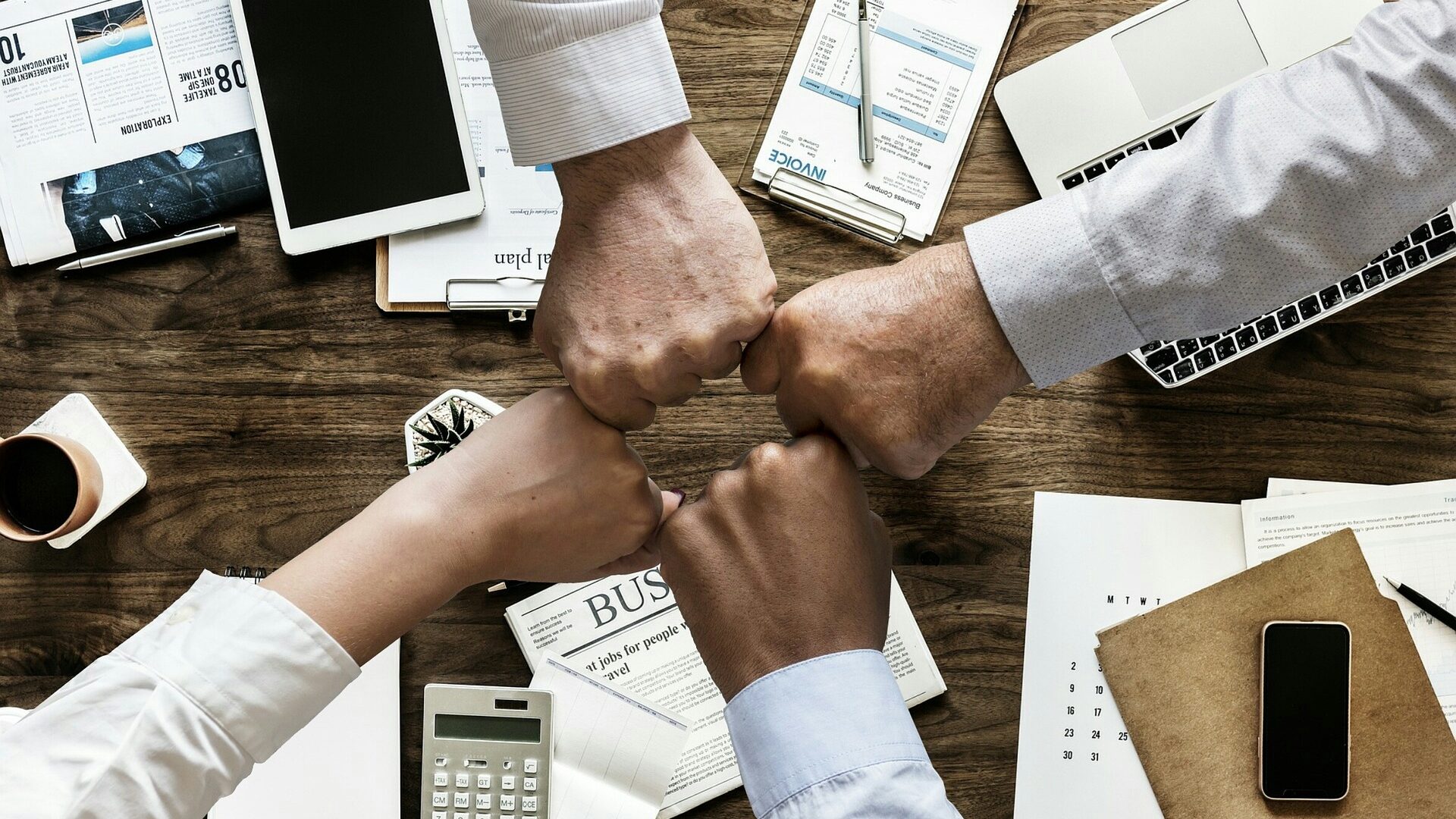 Vier Personen stoßen mit den Fäusten über einem Besprechungstisch zusammen. Laptop, Dokumente und Arbeitsmaterialien symbolisieren Teamarbeit und gemeinsame Zielerreichung.
